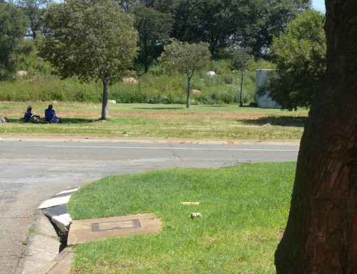 Across the road from the house, loiterers sit in the shade of a tree.